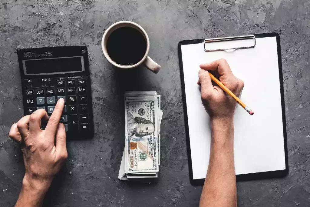 Hands calculating costs on calculator, writing on clipboard, and cash on desk. AFSL application costs.
