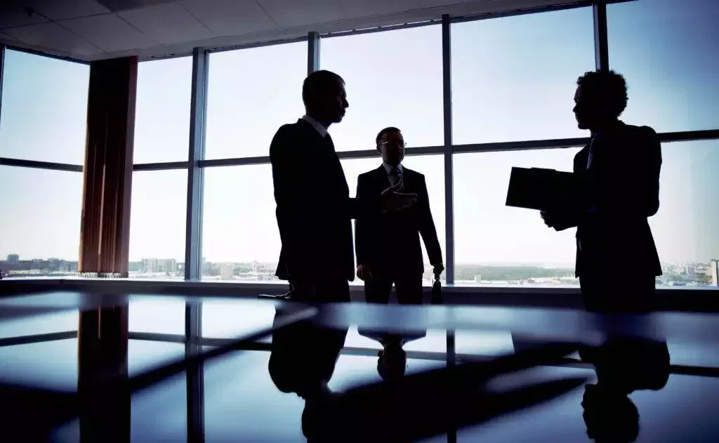 Three businessmen in suits discuss business near a large window overlooking a city.