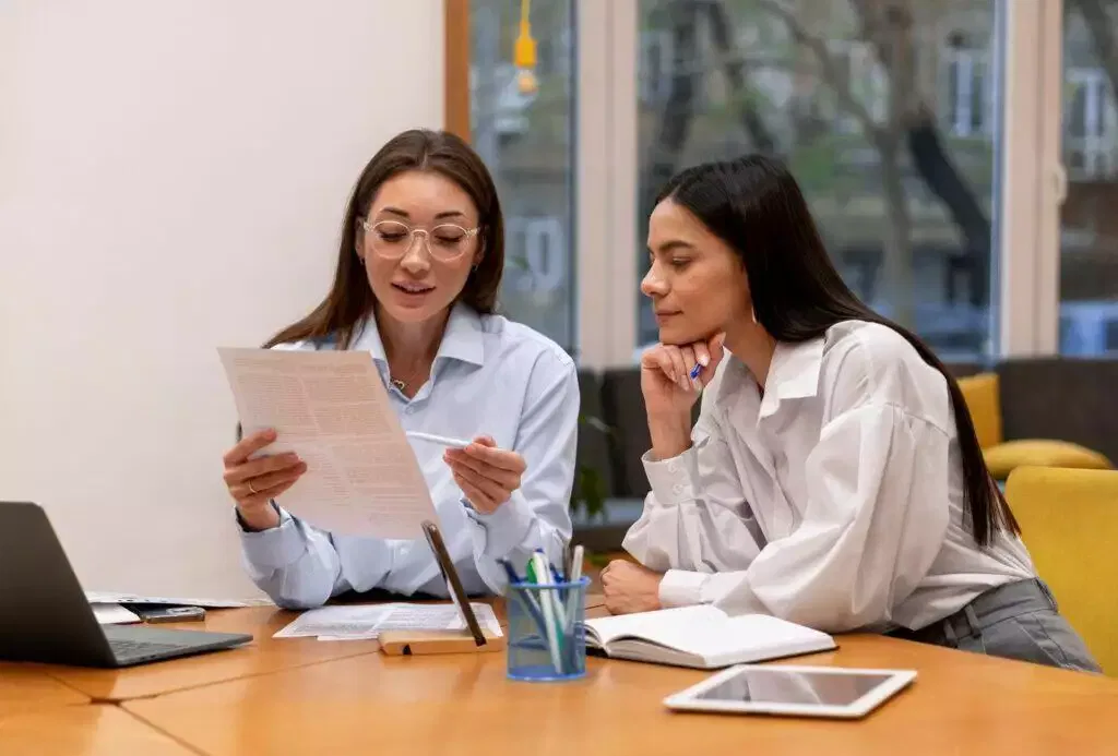 Two businesswomen review documents at a desk, discussing financial matters.