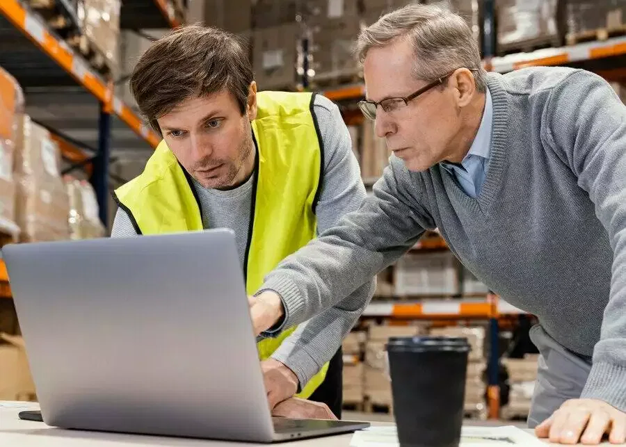 Two men review a laptop in a warehouse. One man wears a safety vest.