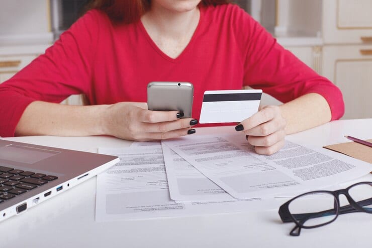 Woman reviewing financial documents, using smartphone and credit card.