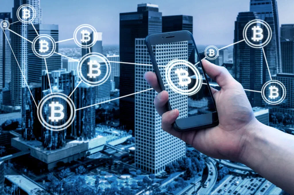 Hand holding a phone with Bitcoin icons overlaid on a city skyline. Digital assets concept.