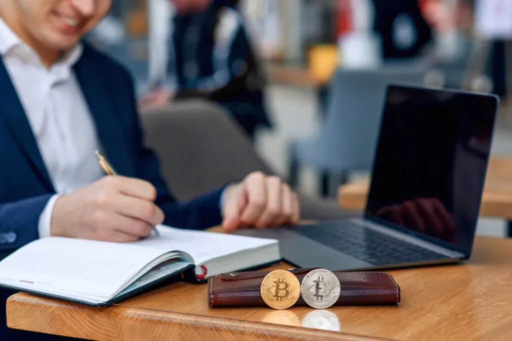 Man writing in notebook with laptop and Bitcoin, Litecoin coins on the table. Fintech concept