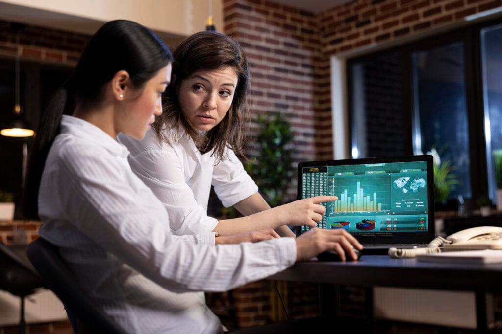 Two women analyze financial data charts on a laptop. Fintech BIN sponsorship analysis.