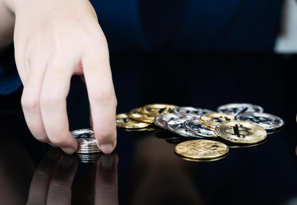 Hand stacking silver Bitcoin coins, with gold and silver crypto coins scattered on a glossy surface.