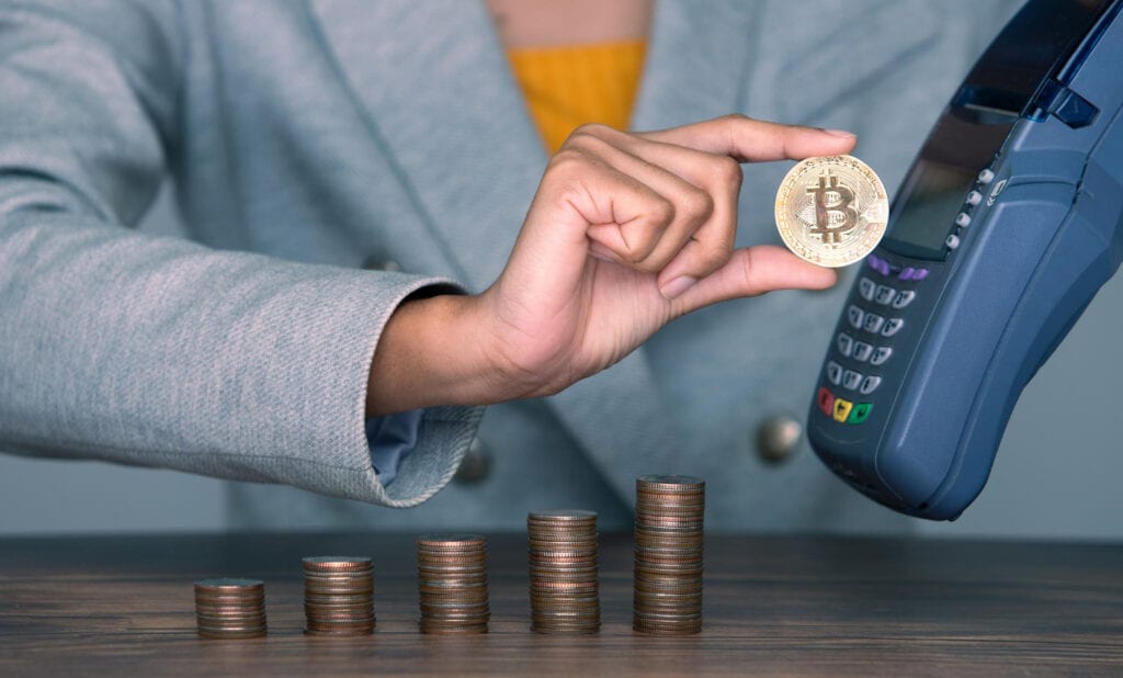 Hand holding Bitcoin near a card reader, with stacks of coins representing financial growth.