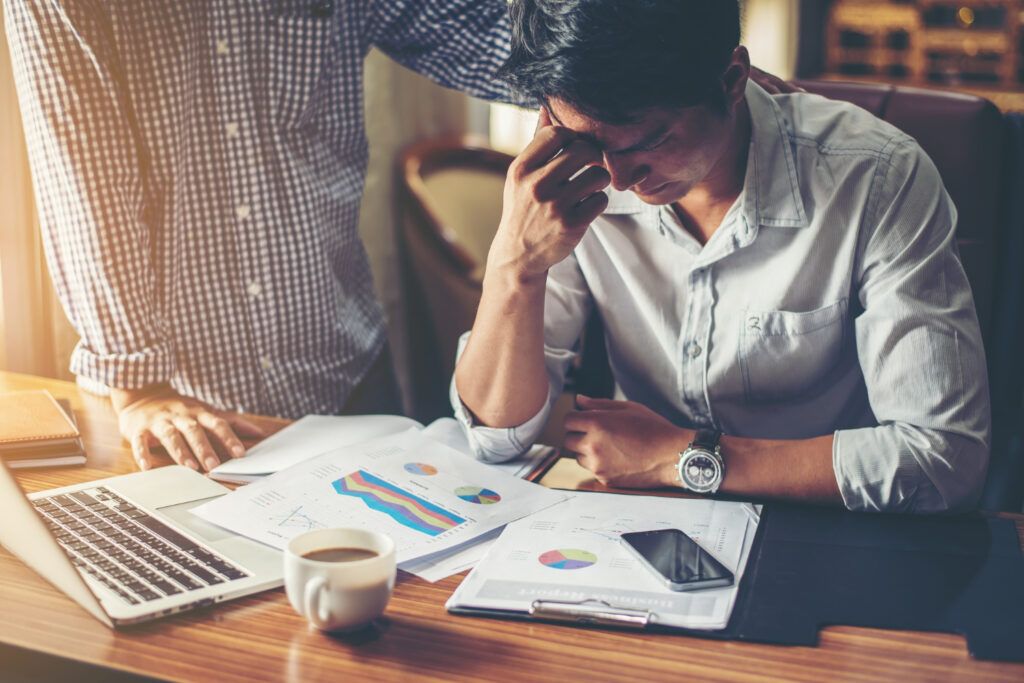 Stressed man at desk with financial reports, being consoled. Financial hardship compliance.