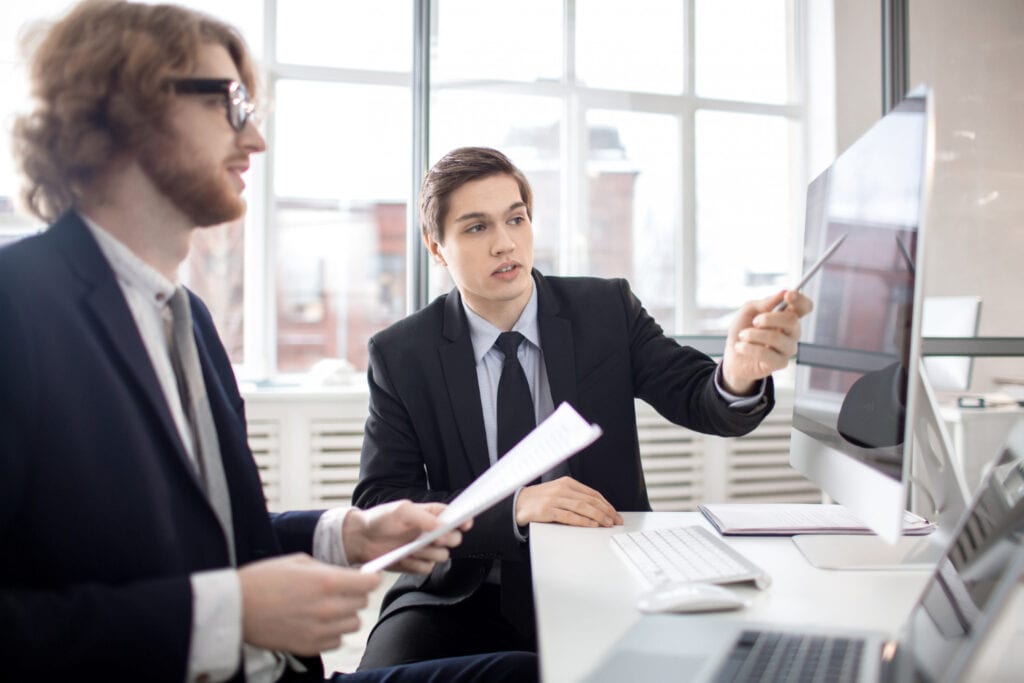 Two men in suits discuss ASIC compliance risks for CFD brokers at a desk.