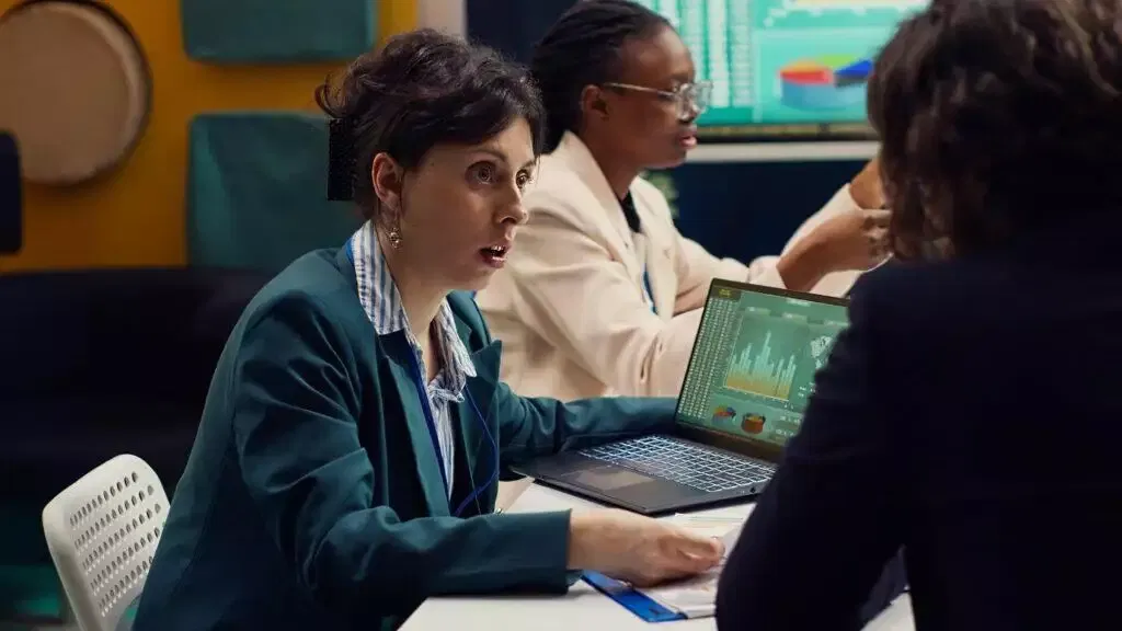 Businesswomen reviewing financial data on laptop, discussing CFD trading strategies.