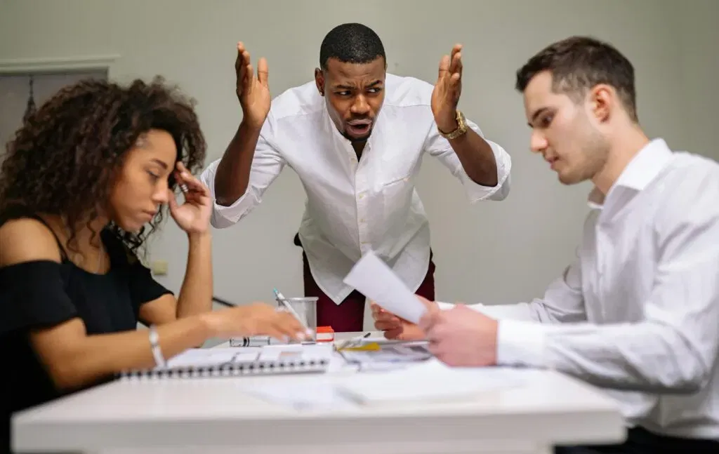 Three colleagues in a tense internal dispute resolution process, discussing complaints at a table.