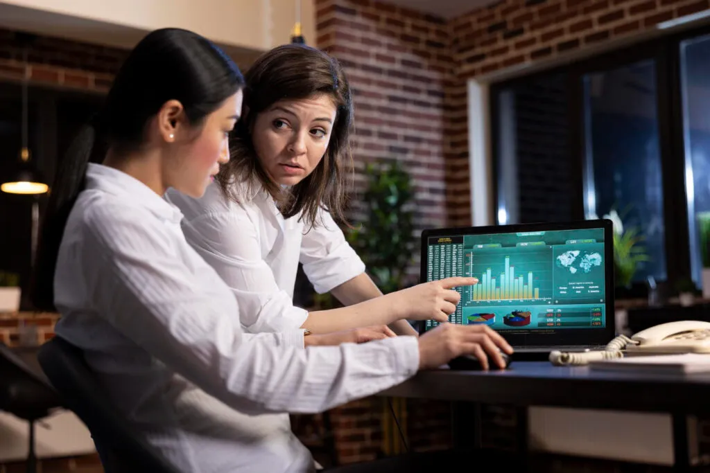 Two women analyze financial data charts on a laptop. Fintech BIN sponsorship analysis.