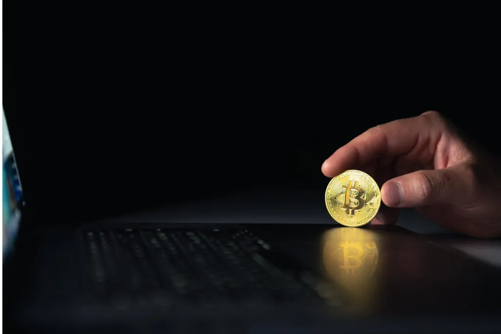 Hand holding Bitcoin over laptop, reflecting on screen. Digital asset concept.