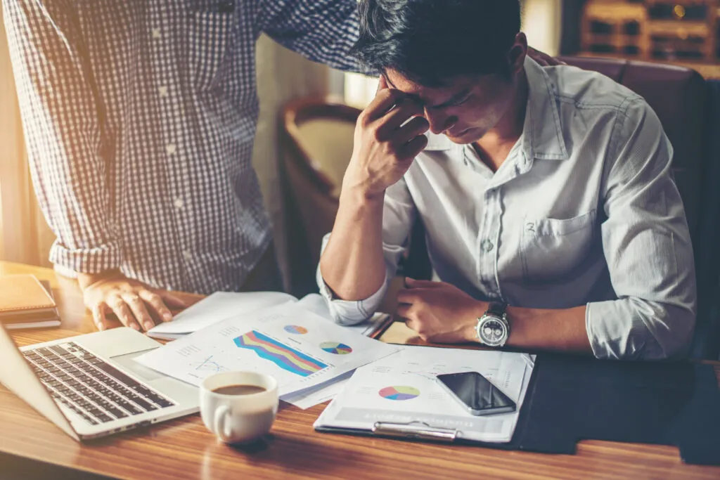 Stressed man at desk with financial reports, being consoled. Financial hardship compliance.